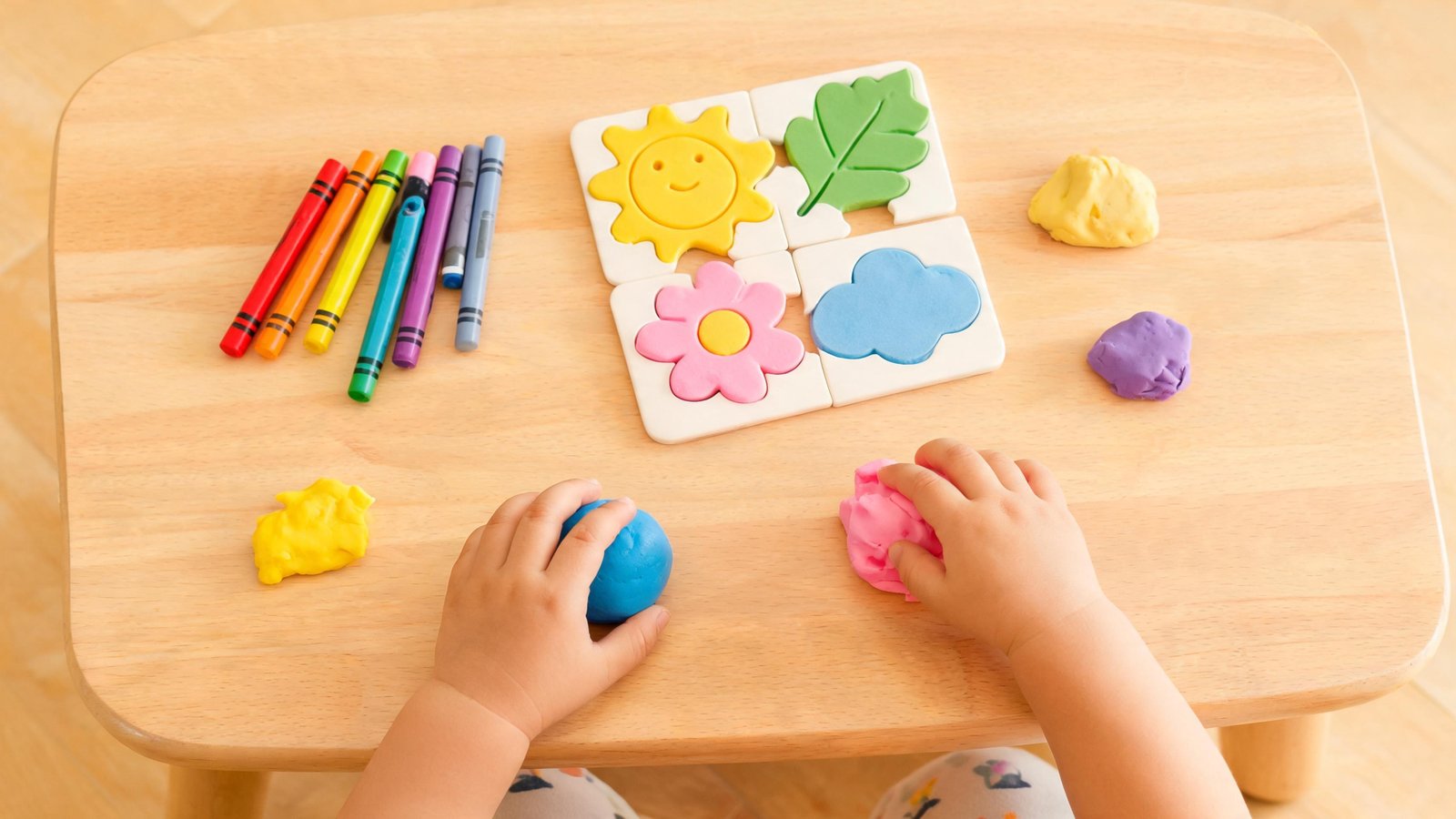 Illustrative image: a child's hands with play dough, crayons and a puzzle on a small table.